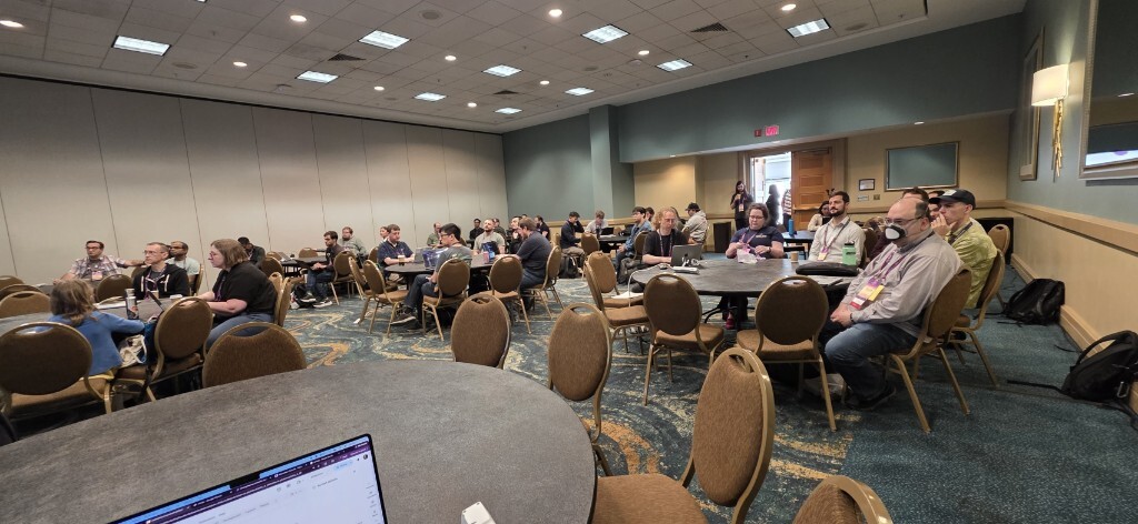 A wide view of the PyCon US Maintainer Summit in Long Beach, with dozens of maintainers seated at round tables, many with open laptops, listening to a session.