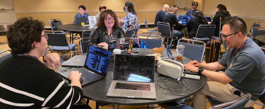 A wide view of a busy conference room during the pyOpenSci sprint at PyCon US 2026 in Long Beach. In the foreground, three sprinters smile and work at a round table with open laptops while many more groups fill the room in the background.