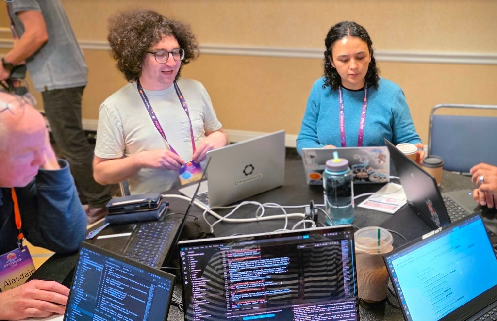 Several PyCon US 2026 attendees work together around a crowded round table during the pyOpenSci sprint. Laptops show code editors and project pages, with purple conference lanyards, charging cables, water bottles, and coffee cups spread across the table.